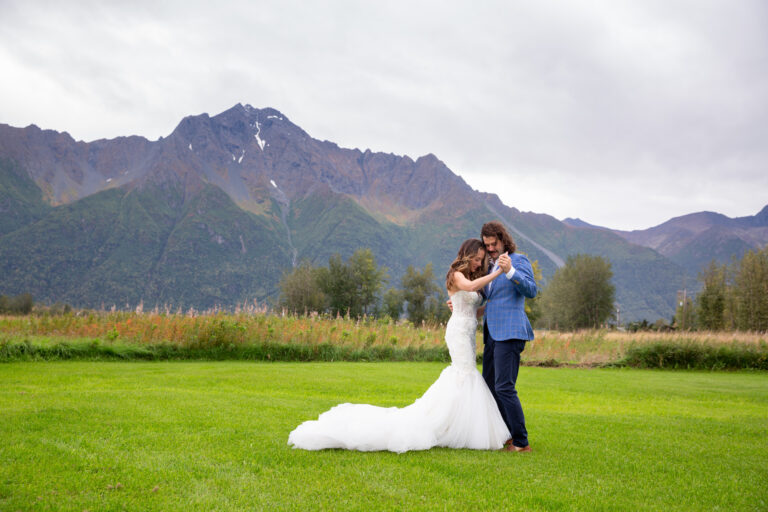 A bride and groom dance together in a bright green grass lawn with mountains behind them.
