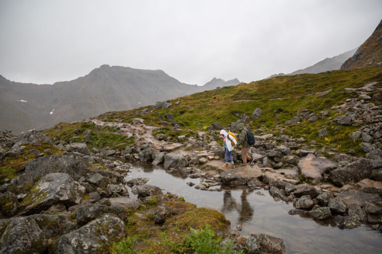 A couple wearing rain jackets, walk hand in hand along a dirt path with a river flowing next to them in Alaska.