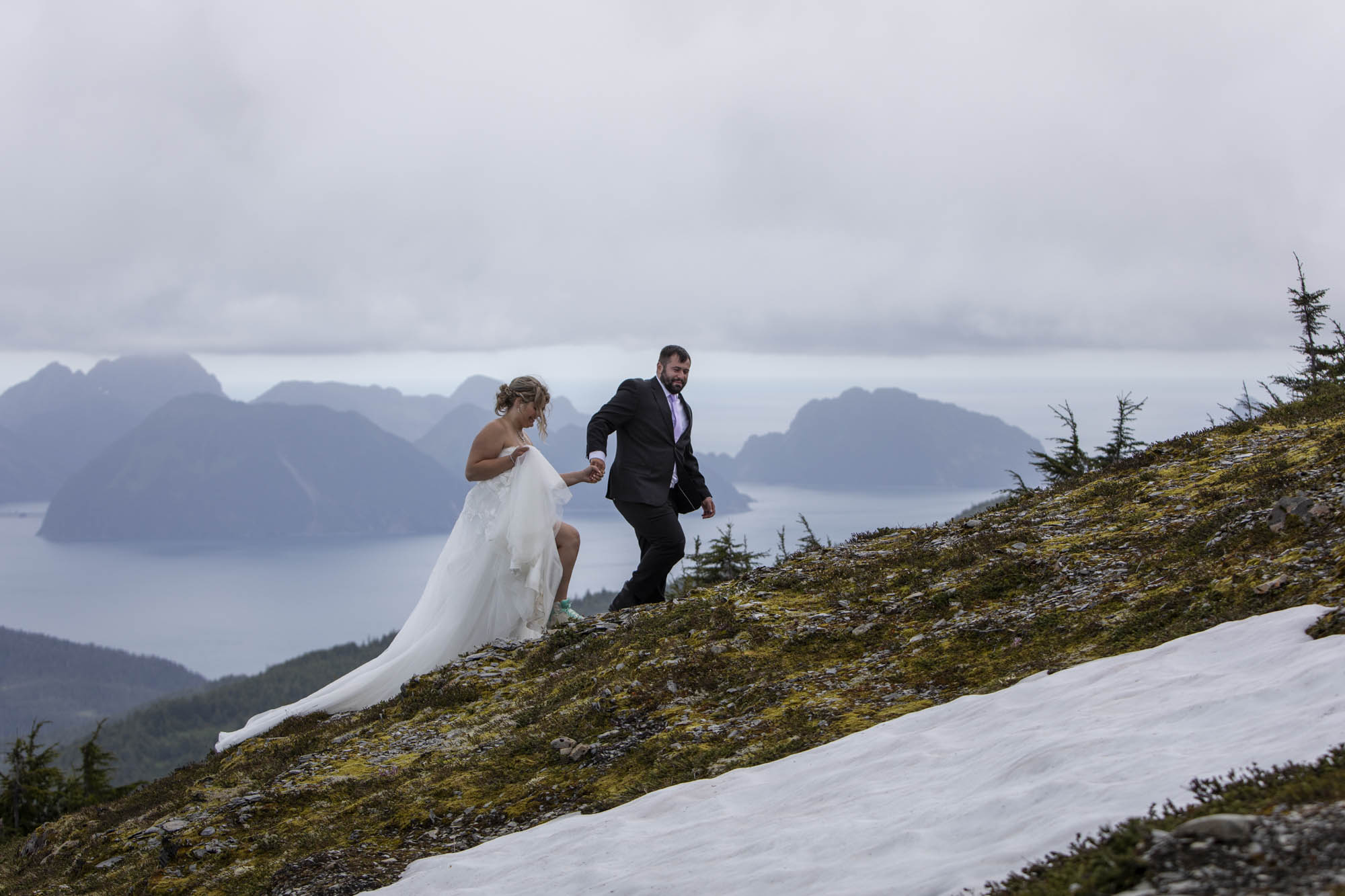 A man in a suit holds hands with a woman in a white wedding dress as they walk up a hill in Alaska after signing their alaska marriage license earlier that day.