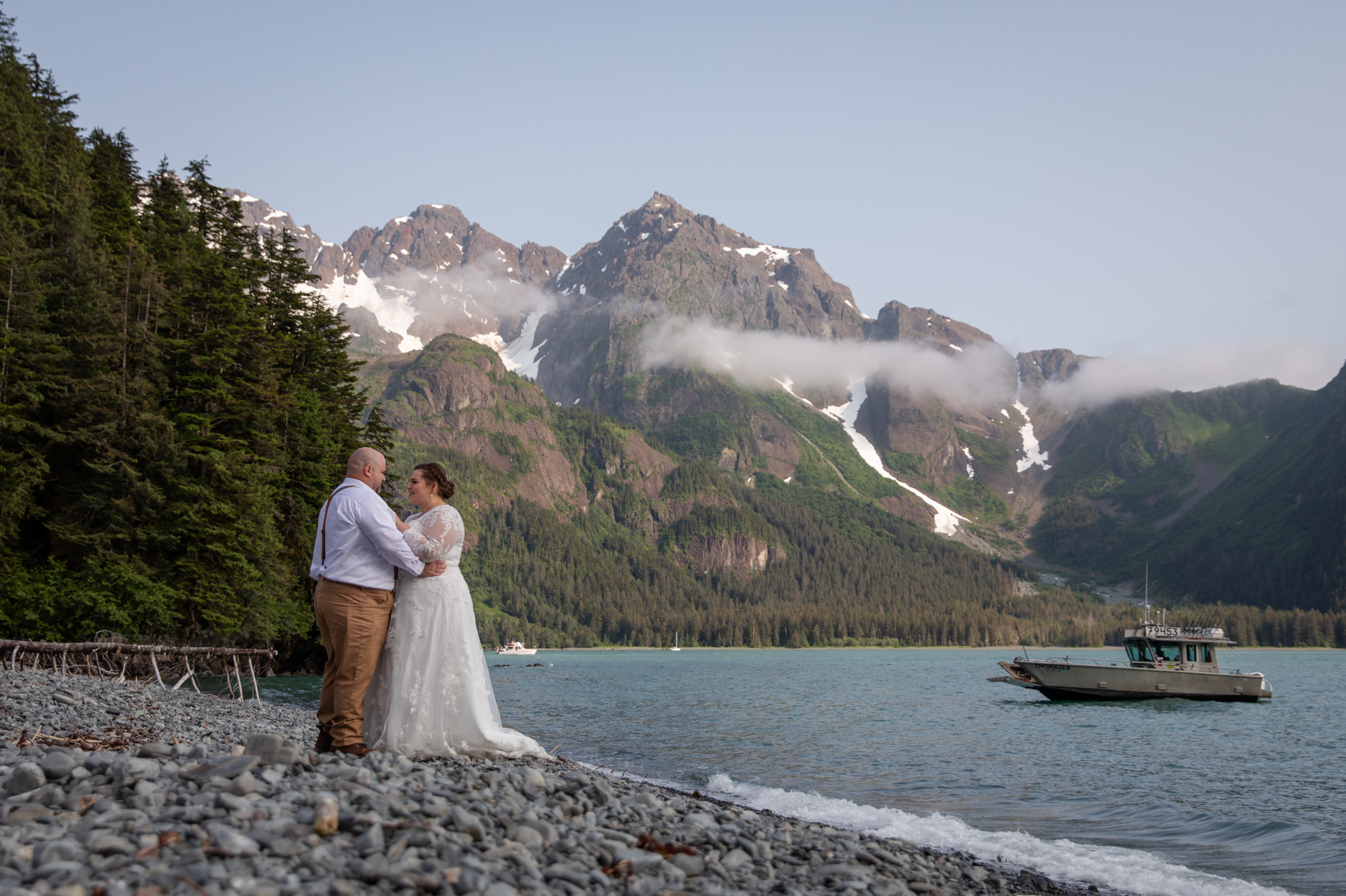 A bride and groom dance with each other on a beach in Alaska while a boat is docked in the water behind them after they signed their alaska marriage license on their wedding day.