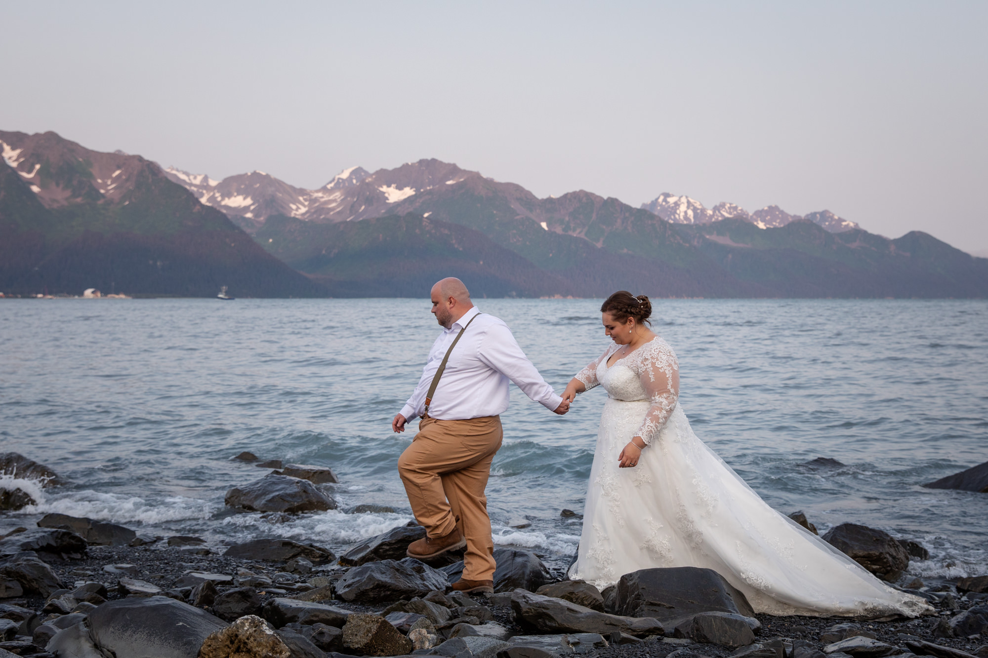 Couple walking along a rocky shoreline in Seward, Alaska with ocean views and snow-capped mountains during a coastal elopement.