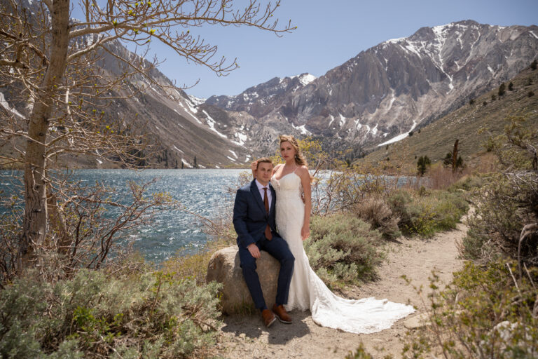 A groom sits on a rock while his wife stands next to him in caressing his head in front of Convict lake in the Eastern Sierra Mountains.