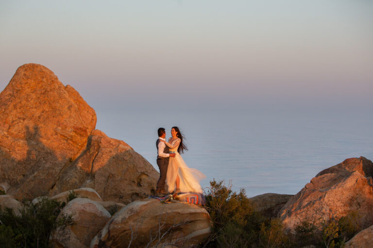 A bride and groom stand hugging each other and smiling as the sunsets around them and their shadow is on a big rock next to them and the ocean behind them