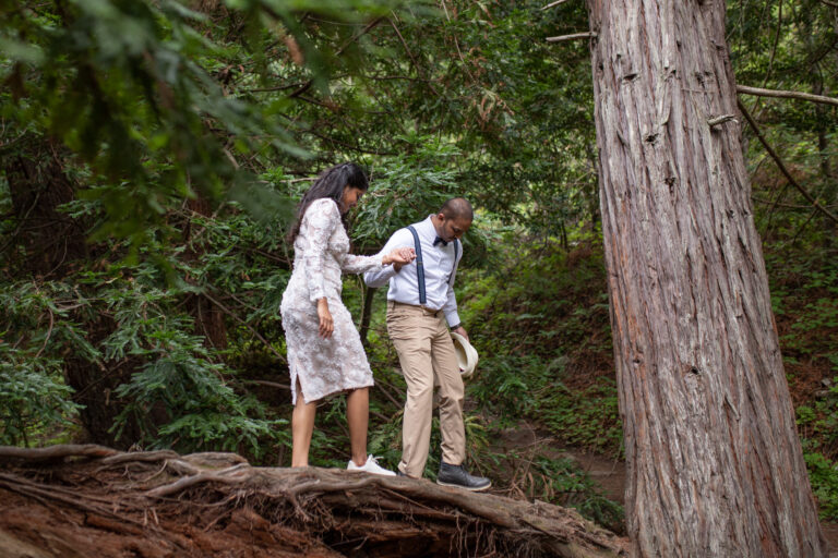A groom walks along a fallen tree in Big Sur, holding his bride's hand and leading her along the tree.