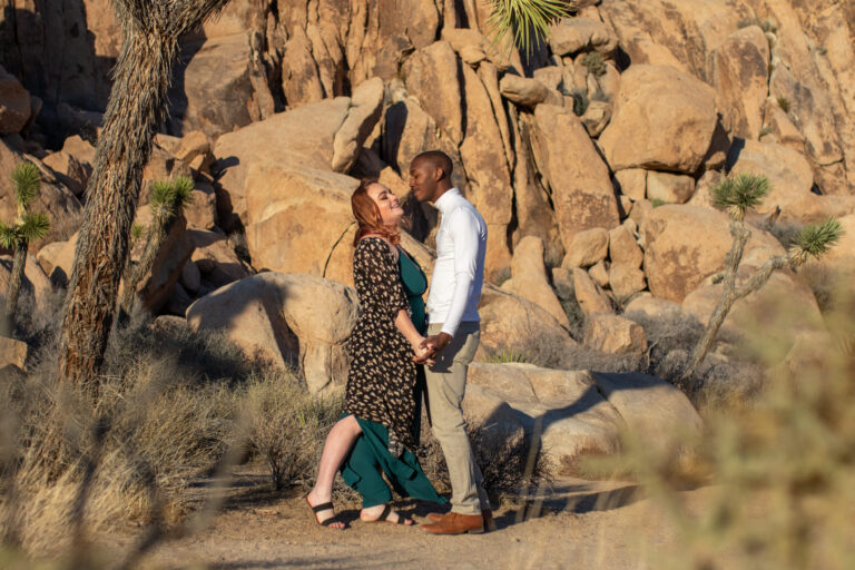 A couple stand facing each other and holding hands in Joshua Tree with boulders behind them