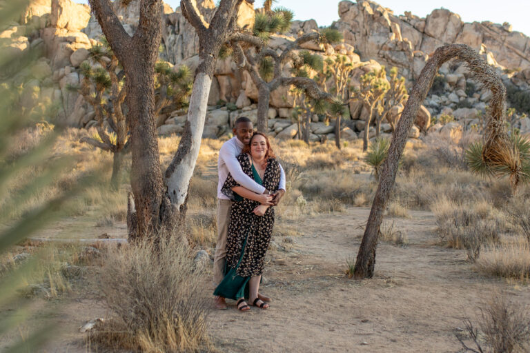 A man stands behind a woman in Joshua tree, wrapping his arms around her waist to keep her warm.