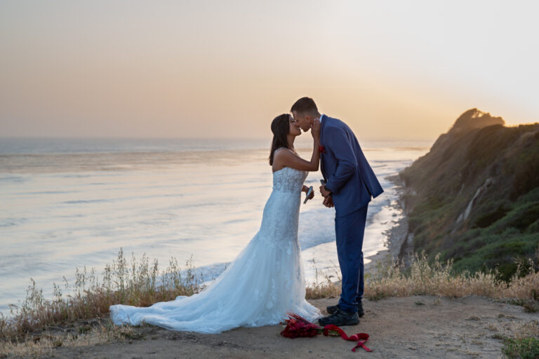A bride places her hand on her groom's face as they kiss with the ocean behind them and a bouquet of flowers at their feet.