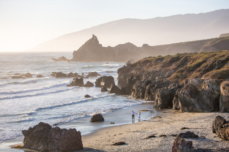 A bride and groom walk along the beach in Big Sur on their elopement day and the rocks stick out of the ocean behind them.