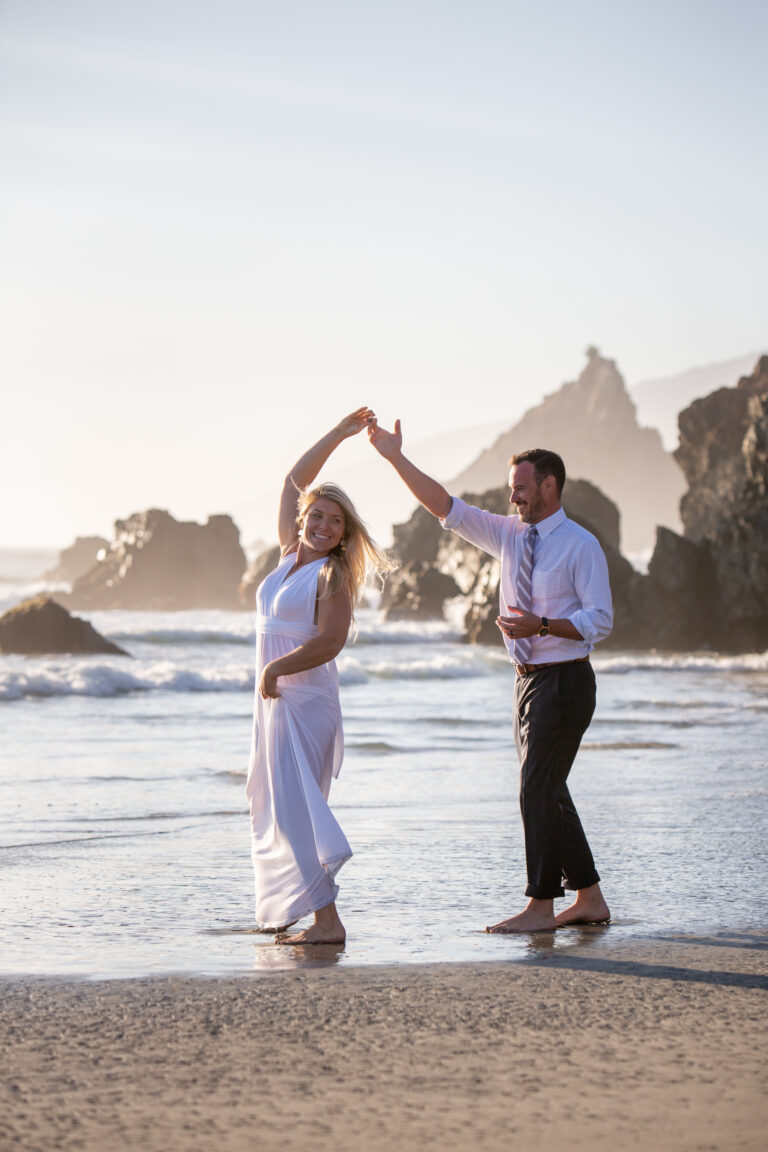 A groom spins his bride as they stand on the beach of Big Sur with giant rocks sticking out of the beach behind them