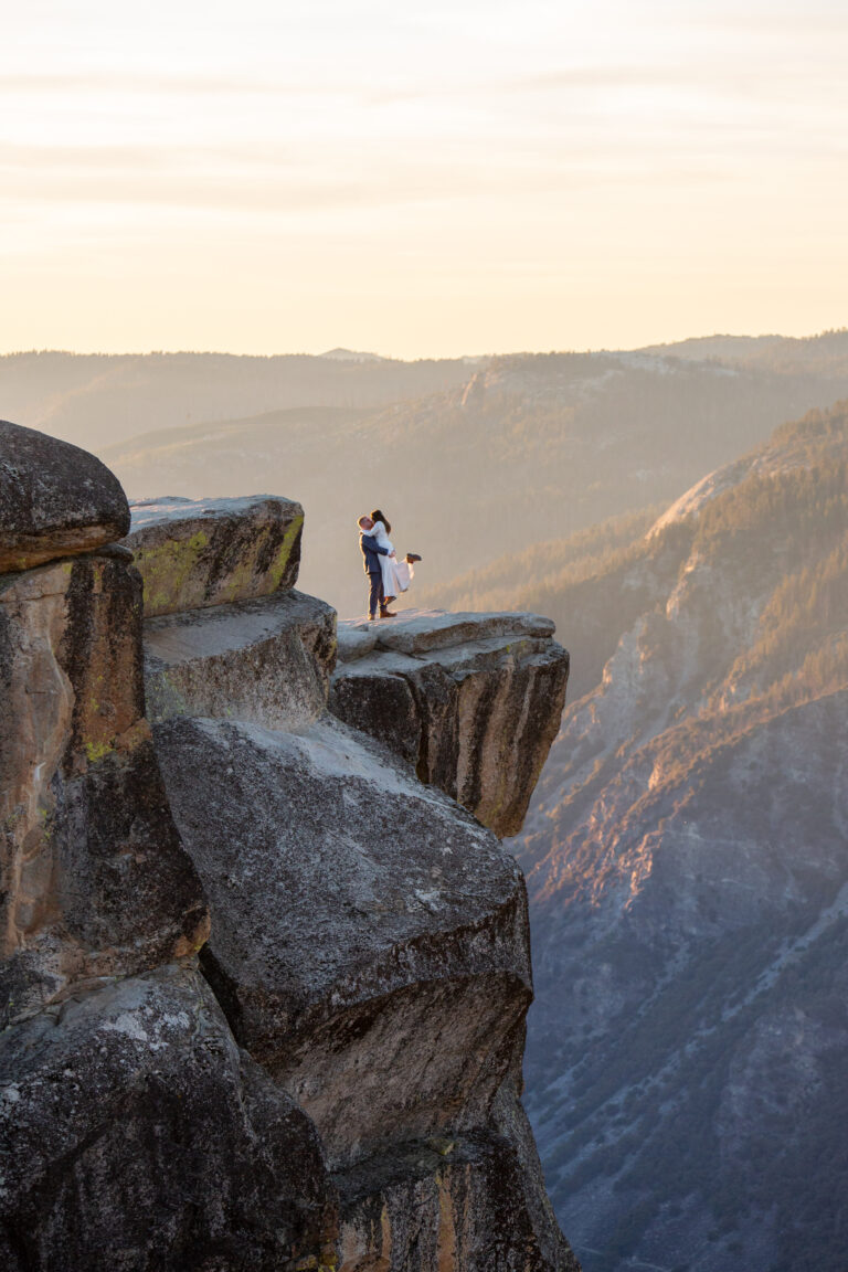 A groom holds his bride in the air as she pops her foot out and the stand on the edge of a mountain cliff in Yosemite at Taft Point.
