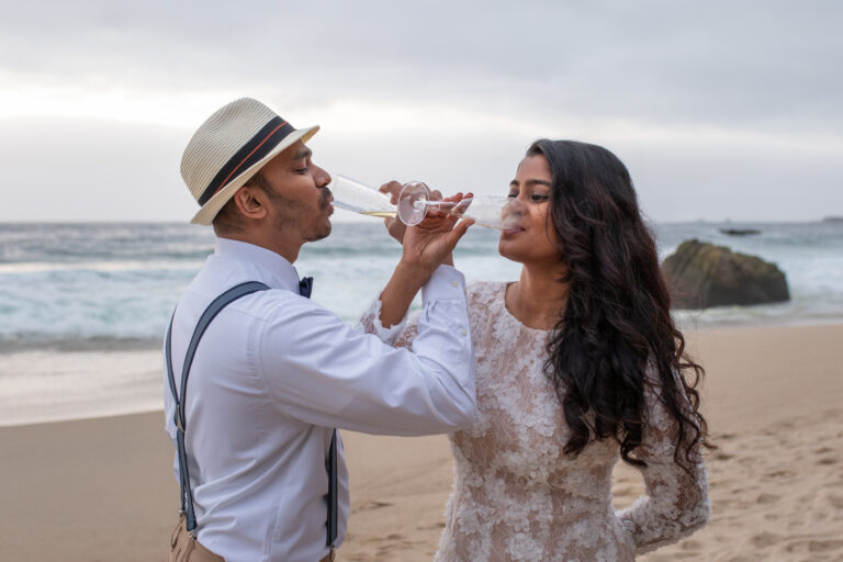 A bride and groom drink out of champagne flutes on their elopement day with their arms wrapped around each others in Big Sur.
