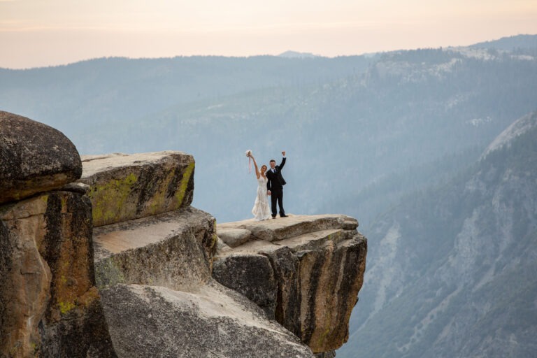 A bride and groom stand with their hands in the air on a cliff in Yosemite National Park
