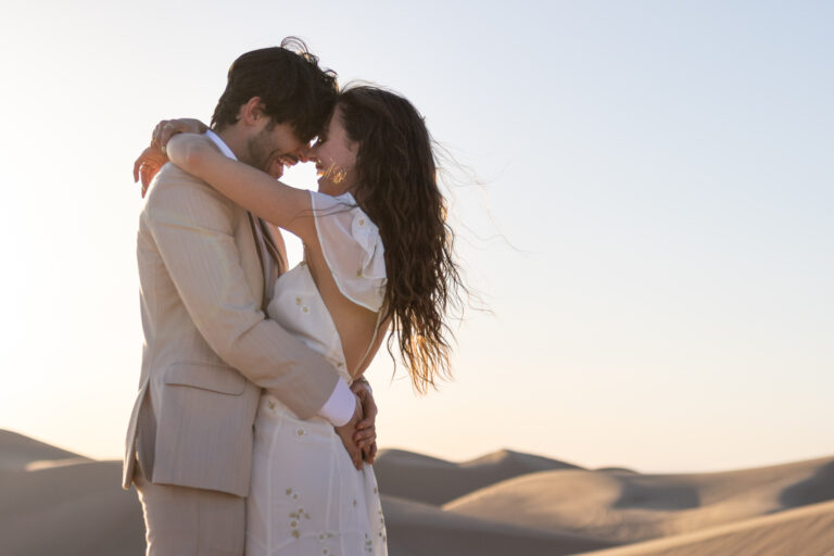 A bride and groom face each other with their foreheads touching as they laugh and stand on a sand dune.