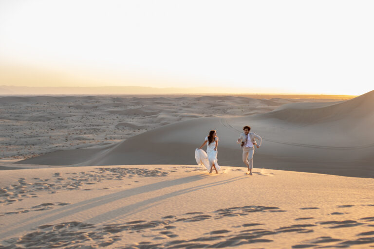 A bride and groom run through the sand with giant sand dunes in the background