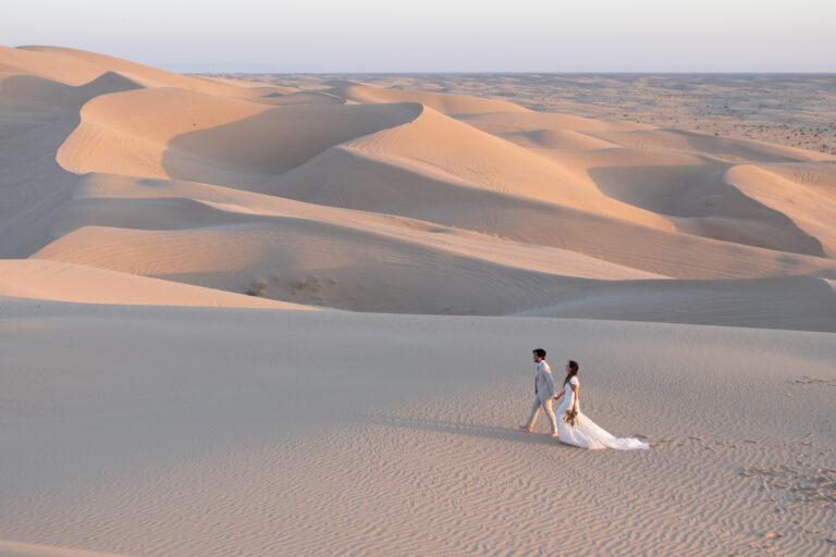 A bride and groom walk hand in hand across a giant sand dune in California as the bride holds her bouquet by her side.