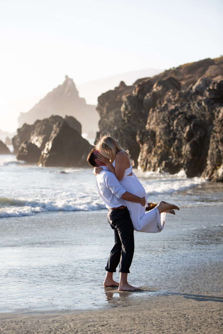 A groom picks his bride up and spins around on the beach in Big Sur as they kiss.