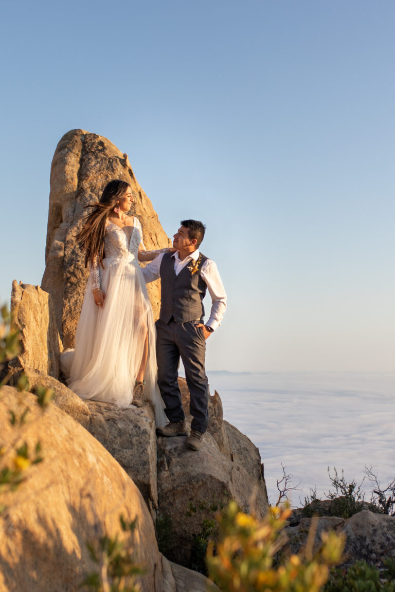 A bride stands on a big rock standing above her groom with her hand on his shoulder and he stands next to her with his hand around her waist.