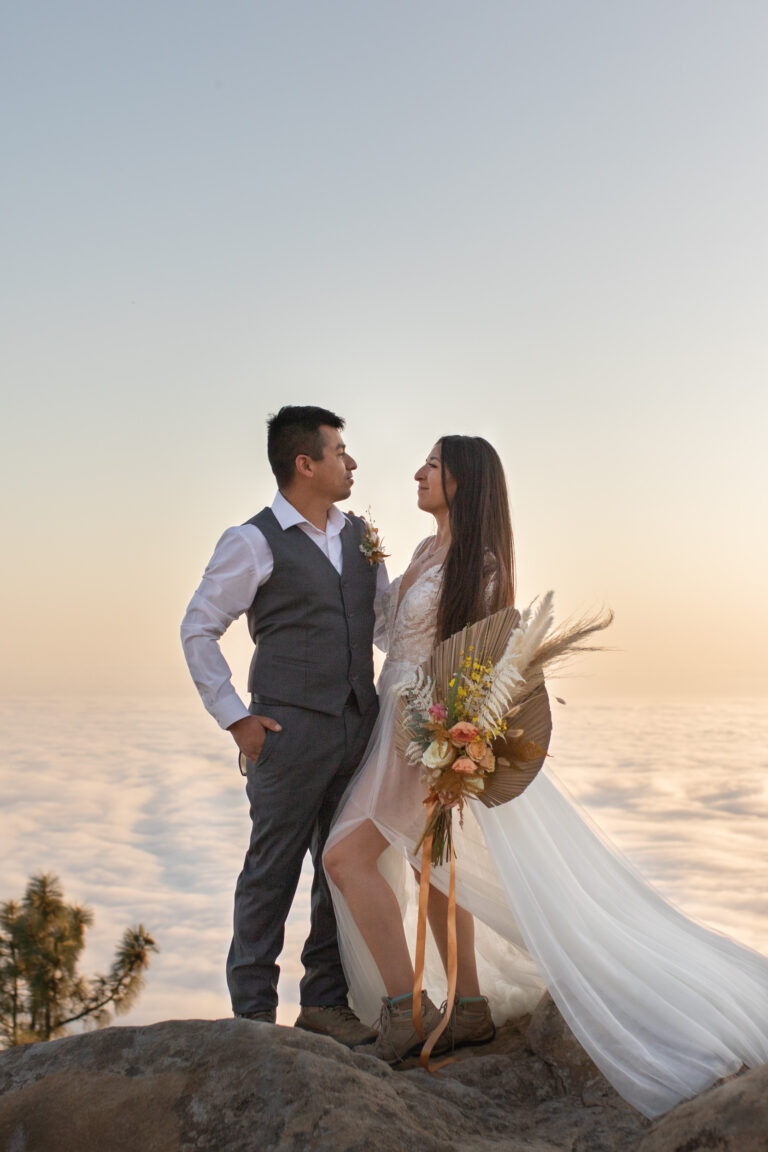 A bride and groom stand facing each other with their arms around each other's waists as a cloud inversion sits behind them and the sunsets.
