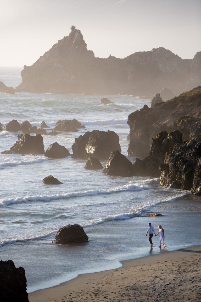 A bride and groom walk hand in hand along the beach in Big Sur and there are big rocks sticking up out of the ocean behind them.