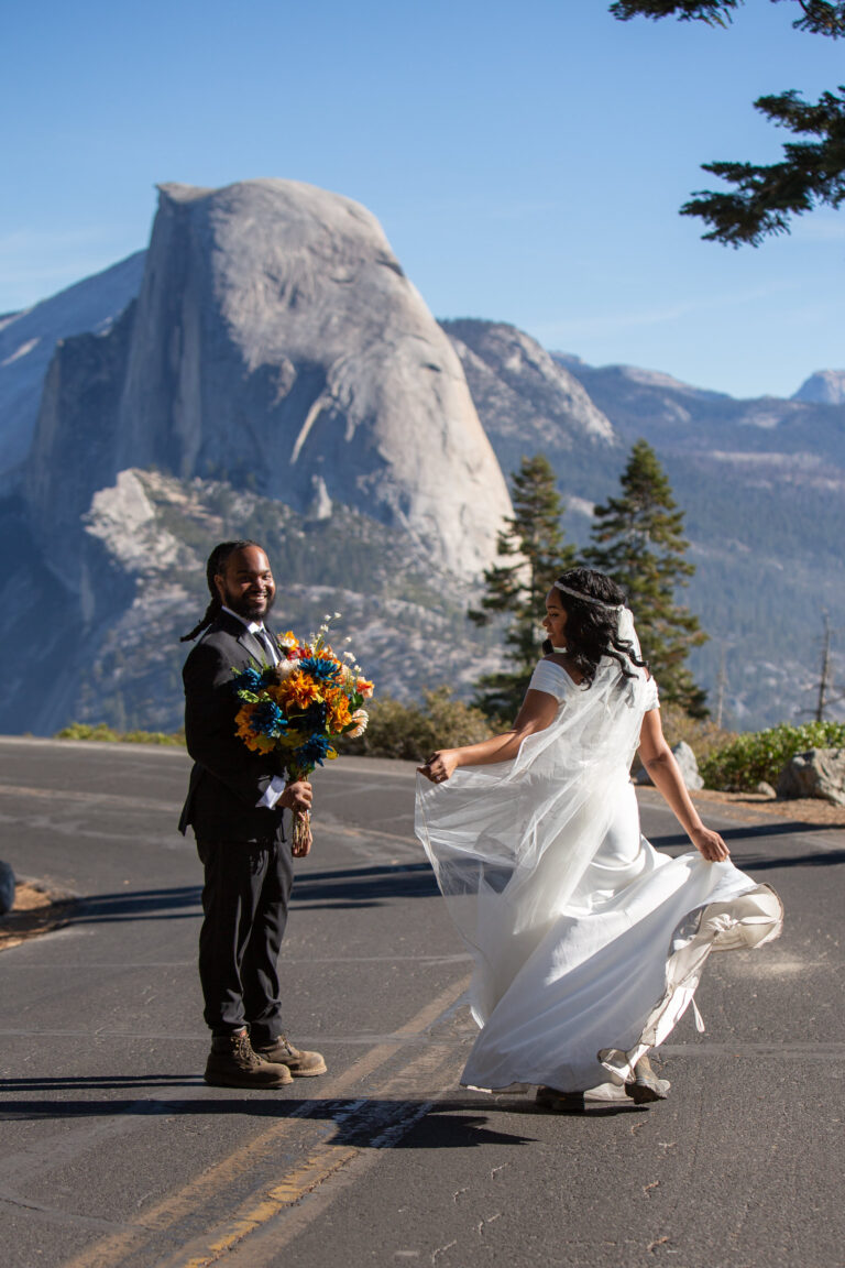 A bride and groom stand in the middle of Glacier Point Road in Yosemite with Half Dome behind them as the groom holds a colorful bouquet of flowers and the bride twirls around.