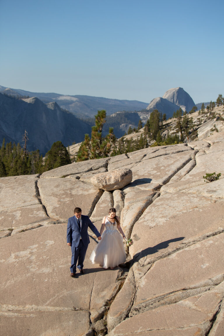 A bride and groom walk hand in hand across a huge granite mountain side with Half Dome showing behind them.