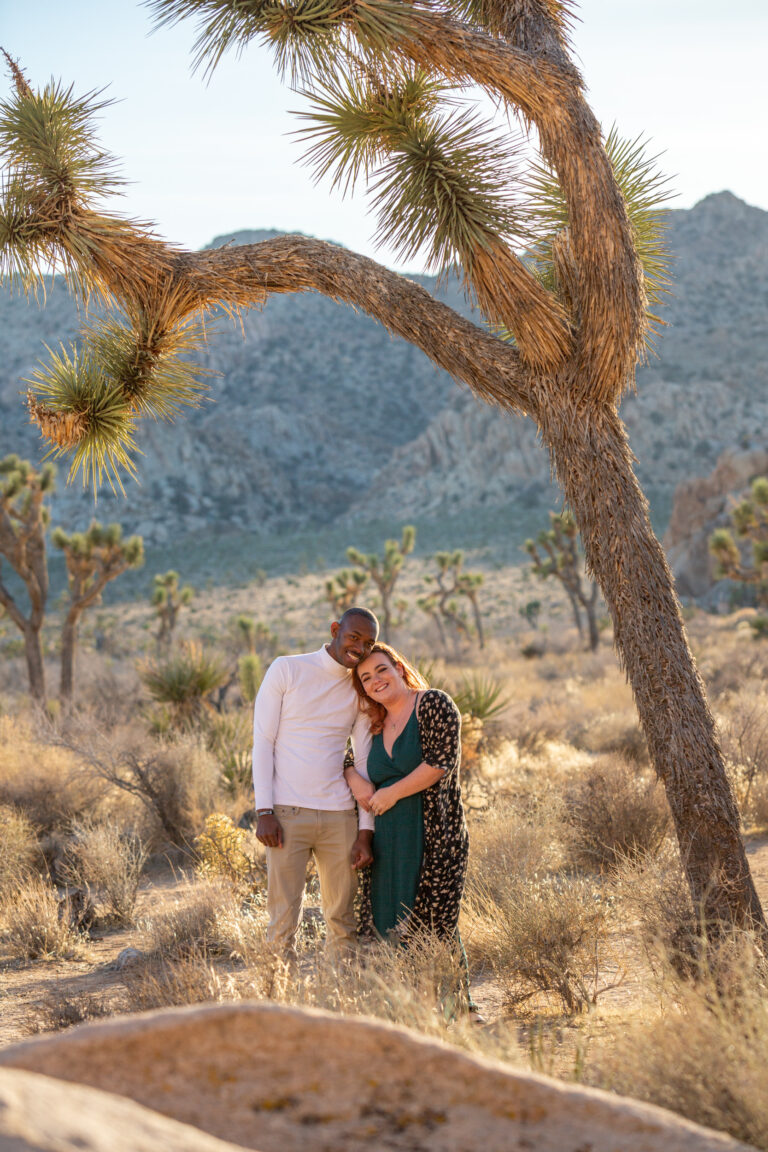 A couple stand next to each other under a Joshua Tree as the woman rests her head on the man's shoulder and loops her arm through his.