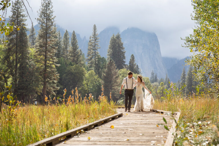 A bride and groom walk along a wooden boardwalk with their arms around each other and the mountains are covered in clouds behind them.