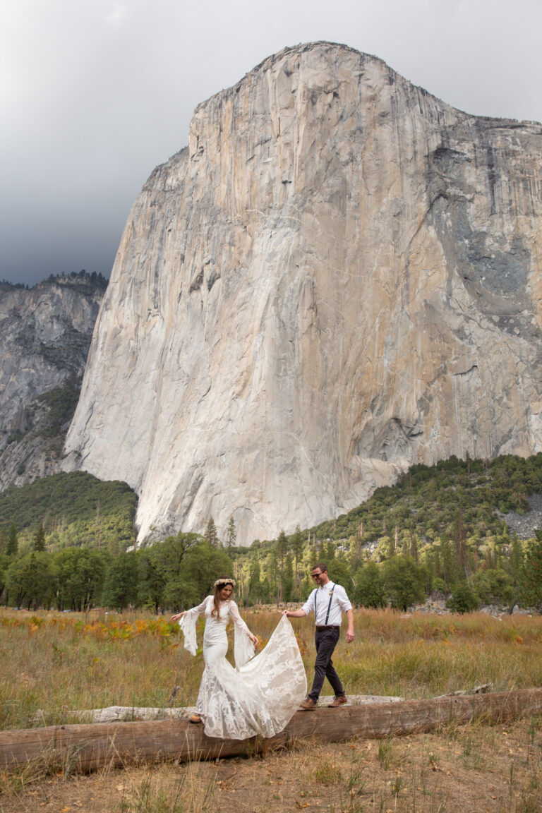A bride and groom walk along a fallen tree in Yosemite National Park with El Capitan behind them as the groom holds up the brides dress behind her.