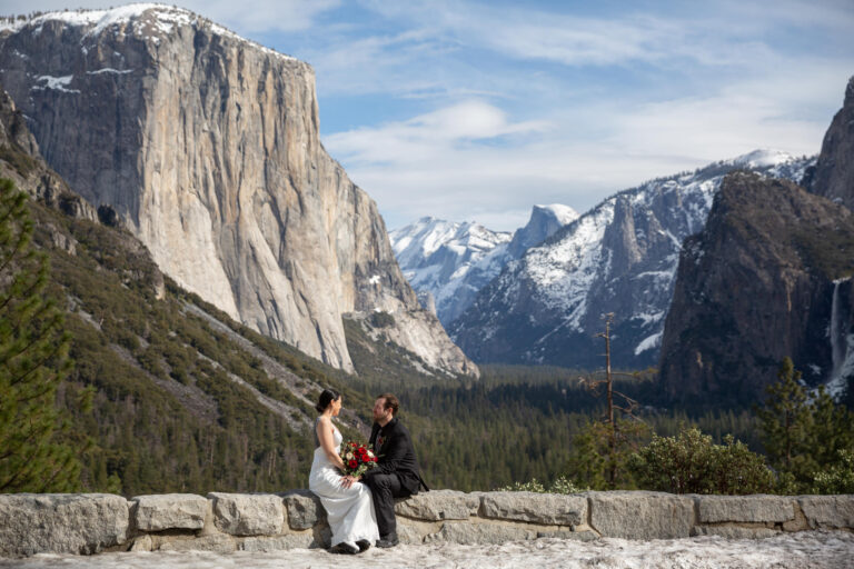 Easily one of the best places in California to elope - here in Yosemite, a bride and groom sit on a rock wall at Tunnel View in Yosemite while the bride holds her red bouquet of flowers and the mountains are covered in snow behind them.