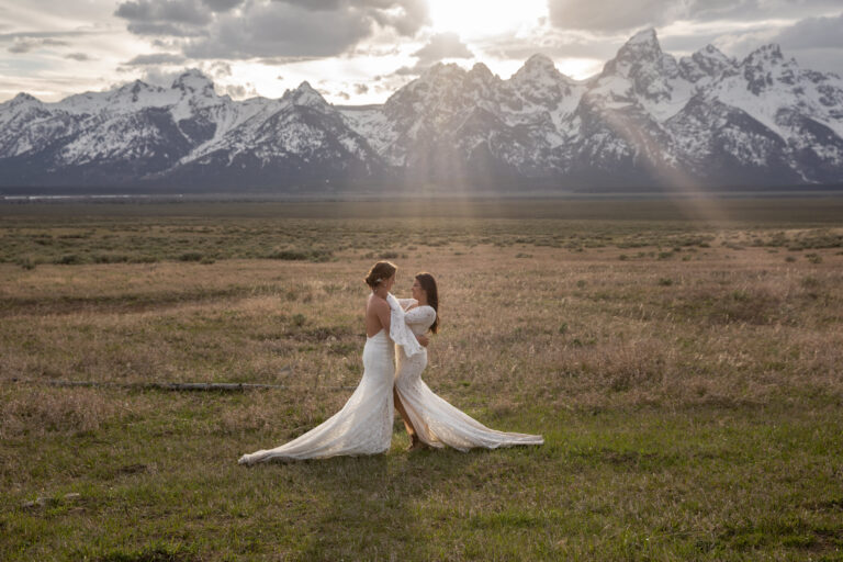 Two brides stand embracing as the sun peeks through the clouds and there are snowcapped mountains behind them