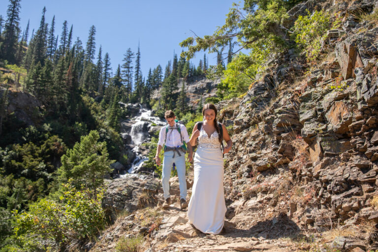 A bride and groom wearing backpacks walk down a rocky trail in Montana as a waterfall flows behind them