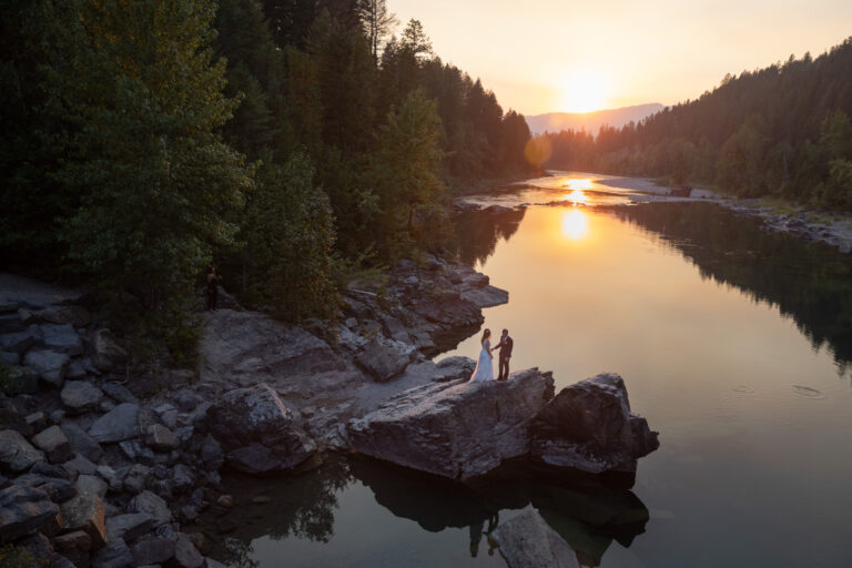 A bride and groom stand on a big rock next to a river as the sunsets behind them
