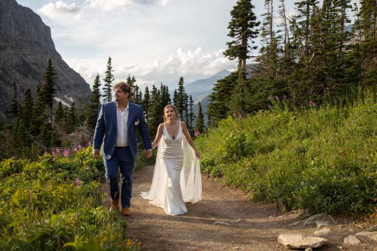 A bride and groom walk towards the camera on a dirt path in Glacier National Park with tall grass and wildflowers blooming around them