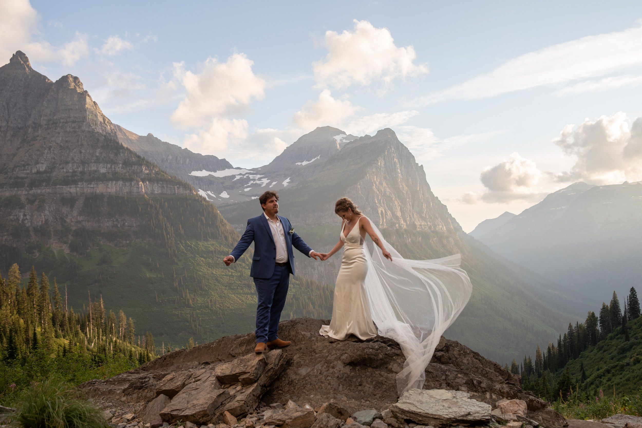 A bride and groom stand on a rock holding hands as the bride's dress blows in the wind next to her in one of the best places to elope in the US