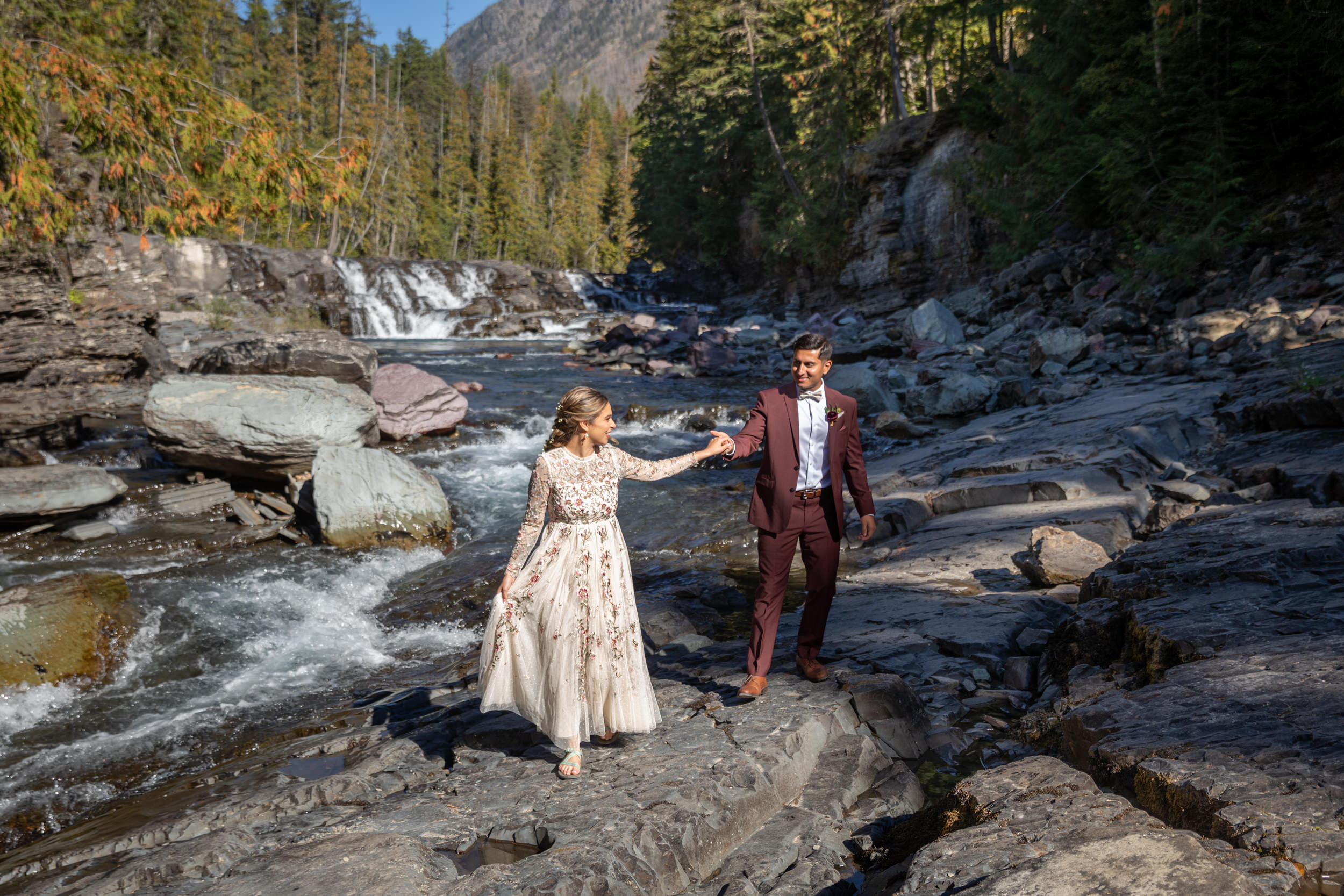 A bride leads her groom by the hand as they walk along rocks next to the river in Glacier National Park on their leave no trace elopement day.