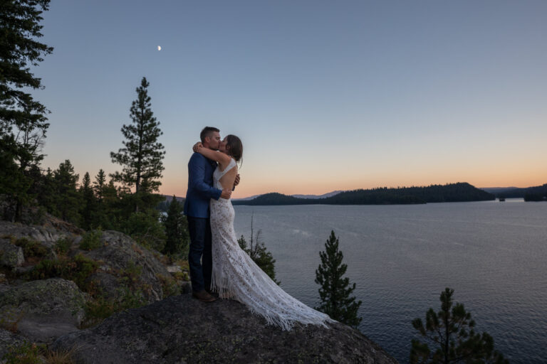 A groom and bride share an embrace and a kiss as the sunsets around them and over the lake they are standing above