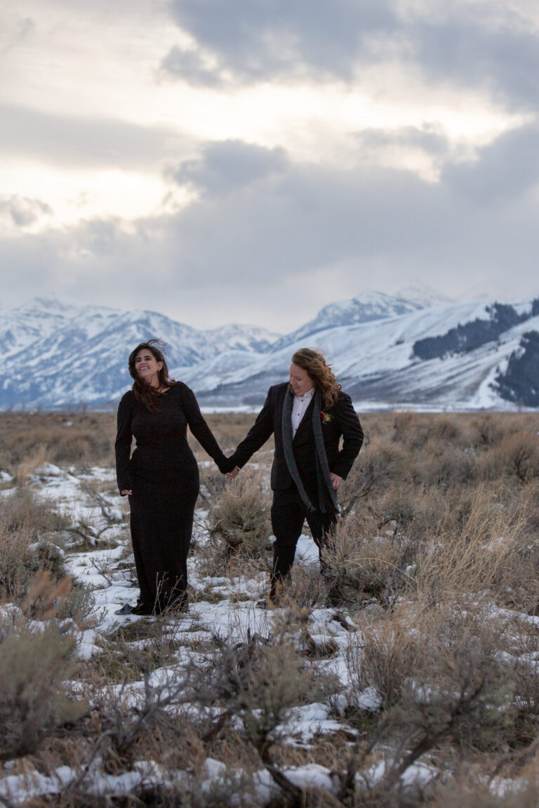 Two brides wearing black stand in a snowy meadow at sunset holding hands