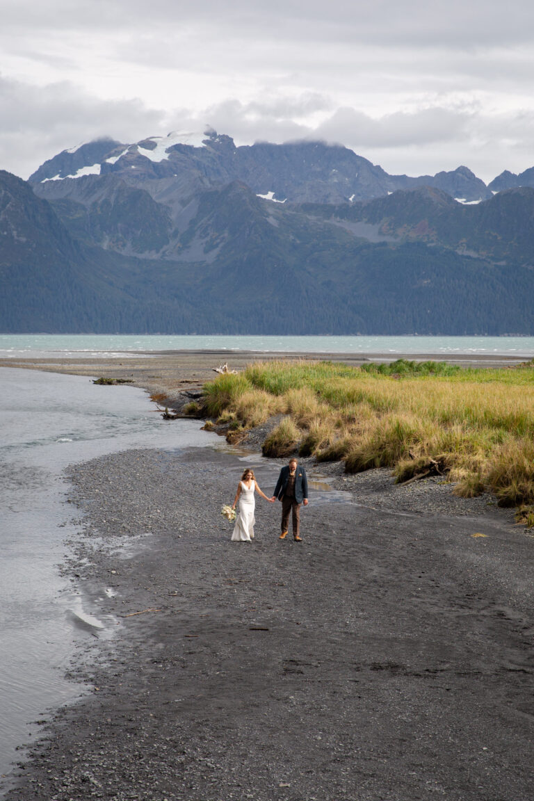 A bride and groom walk hand in hand along a beach in Alaska with mountains towering behind them