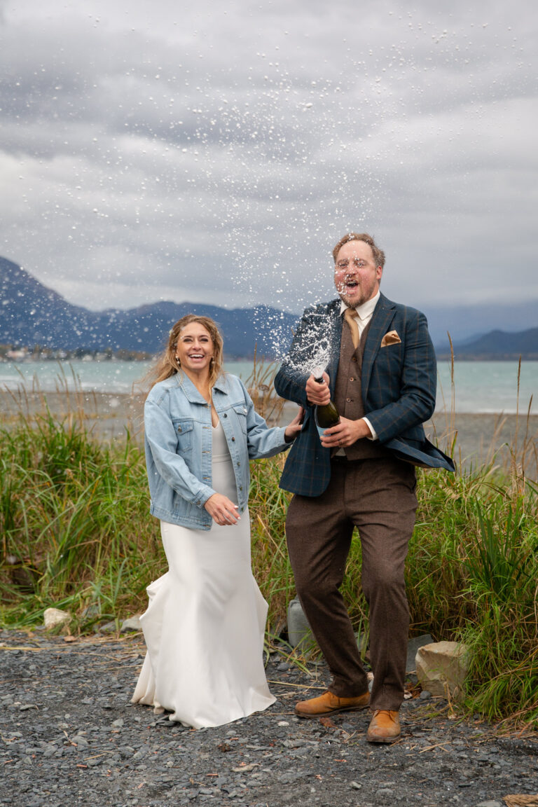 A groom sprays a bottle of champagne at the camera while his bride stands next to him with a hand on his waist.