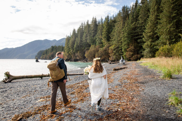 A groom walks with a backpack on and his bride walks next to him along a rocky beach towards a helicopter waiting for them.