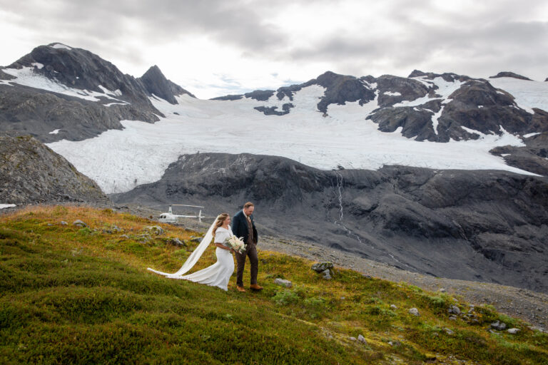 A bride and groom walk hand in hand down a grassy hill with a helicopter and snowy mountain behind them.