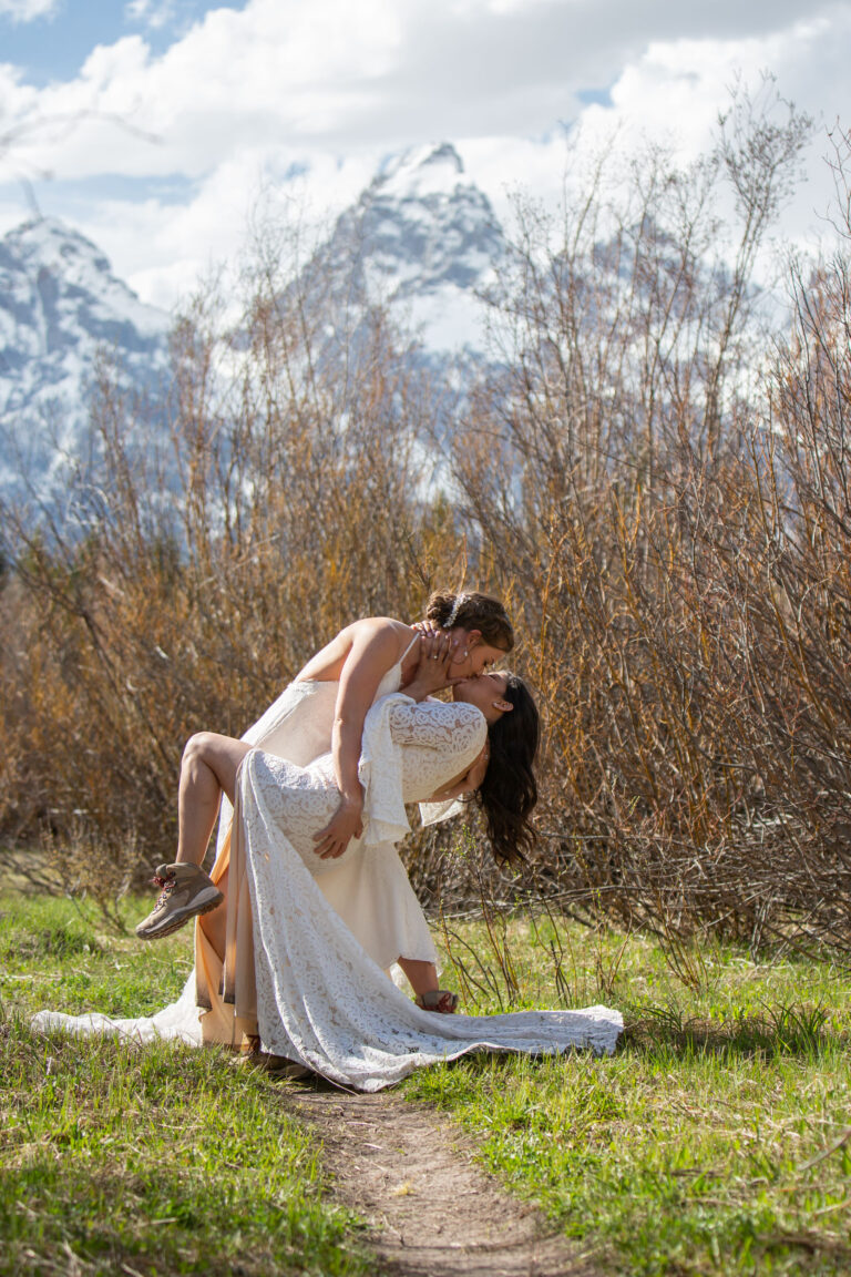 Two brides share a passionate kiss as on dips the other surrounded by high grasses and snowy mountains