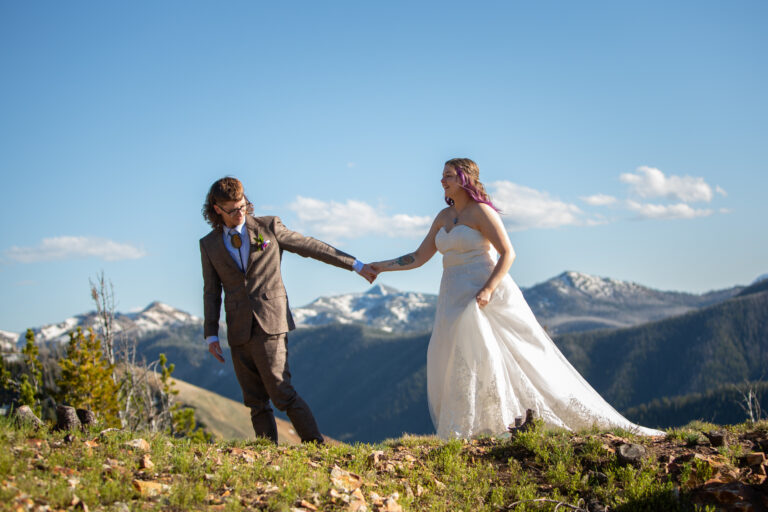 A groom leads his bide by the hand along a hill with snowcapped mountains behind them