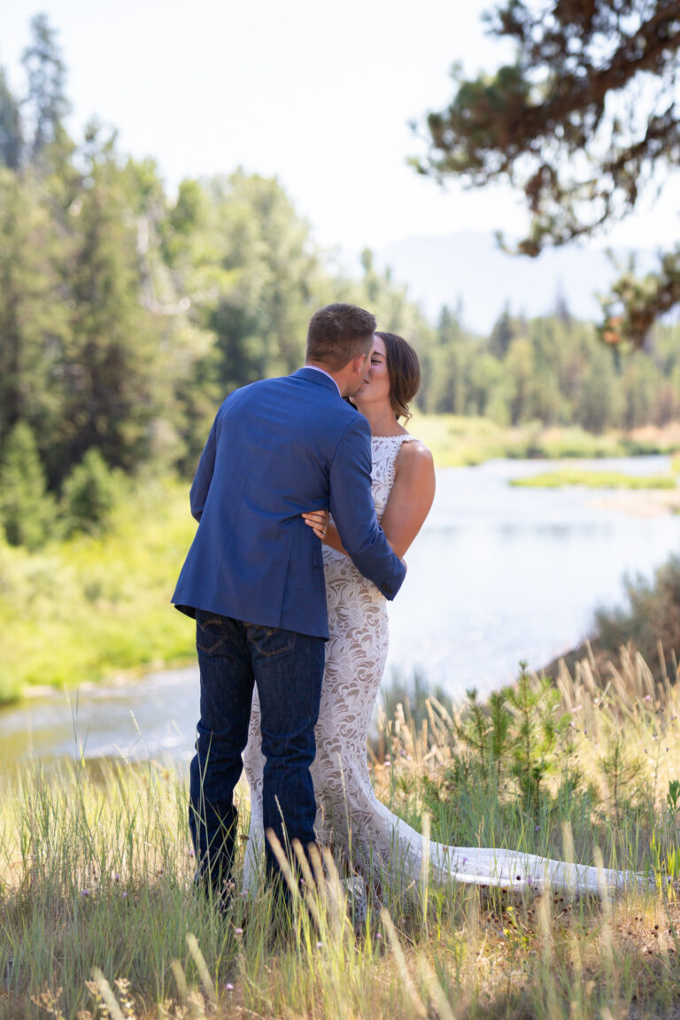 A bride and groom kiss while standing next to a river in a meadow in Idaho