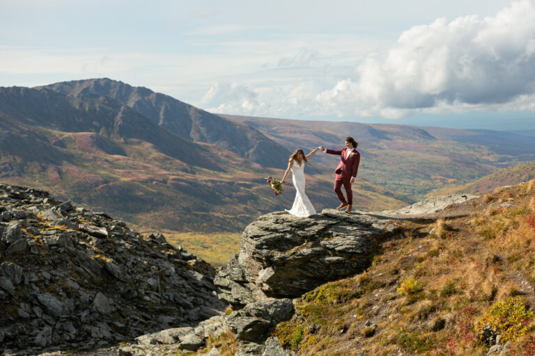 A bride and groom hold hands in the air as they dance on a rock in Alaska on their elopement day.