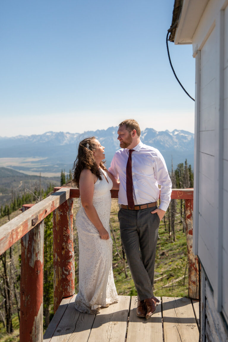 A bride and groom stand looking at each other on a wooden porch of a fire lookout with mountains behind them