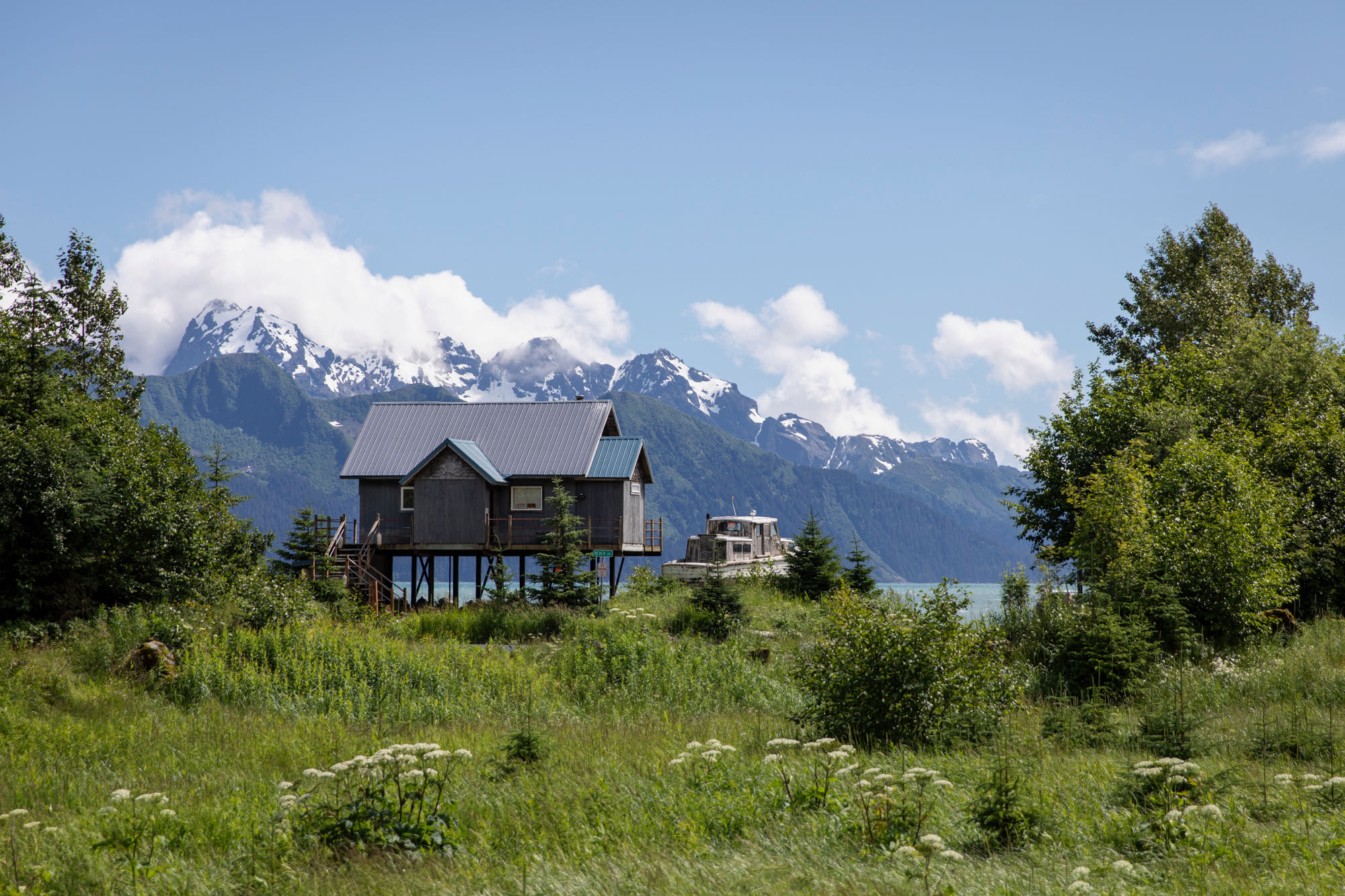 A cabin sits on stilts next to the ocean in Seward, Alaska, with large snow mountain peaks behind it and a grassy green lawn in front of it.