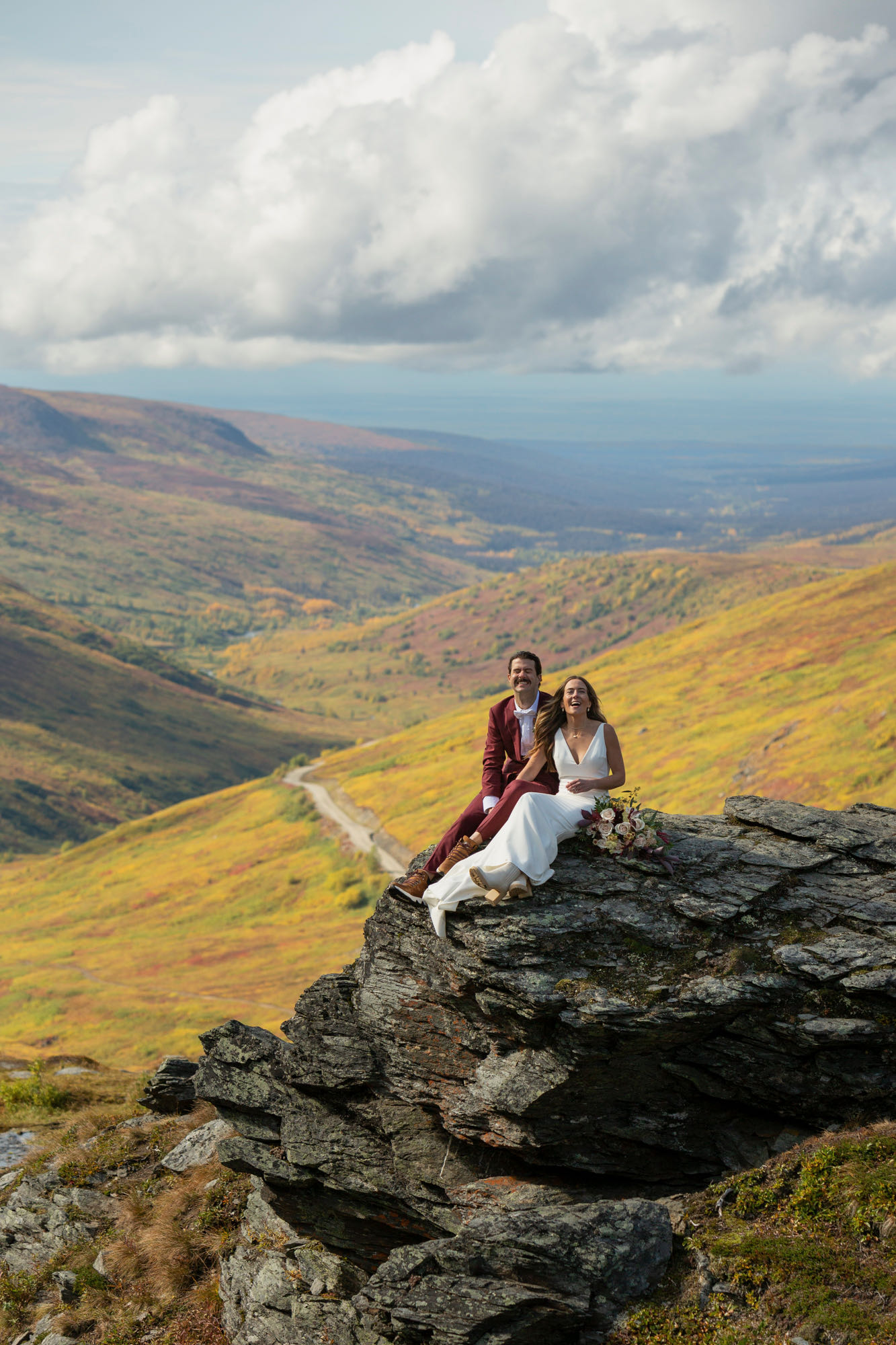 A bride and groom sit on a rock smiling and laughing at the camera after deciding the best time to elope in Alaska was in the fall for them.