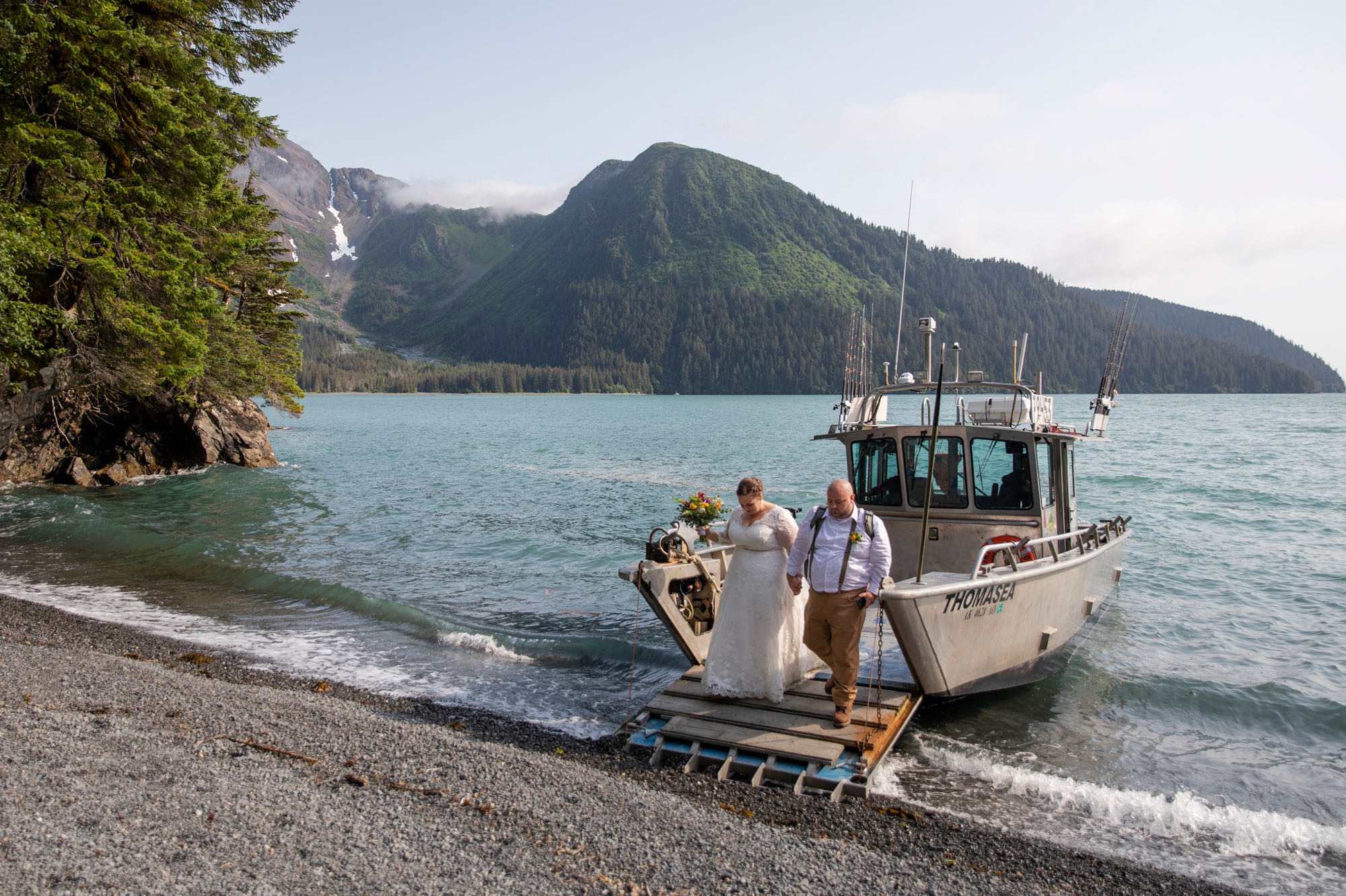 A bride and groom walk hand in hand off a boat as it anchors on the shore of a rock beach in Alaska on their wedding day. 