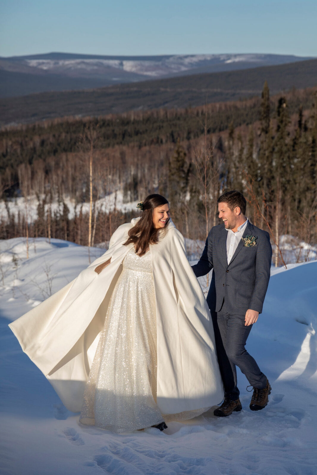 A bride twirls her dress and cape as her groom stands next to her smiling and laughing at her. 